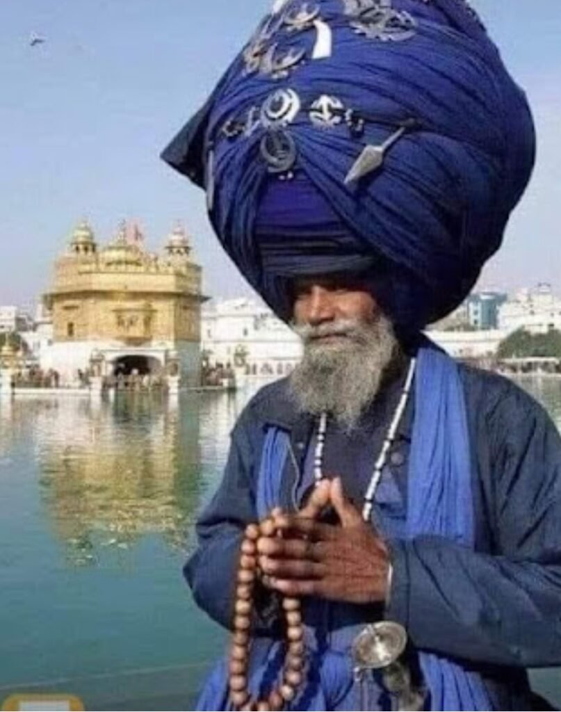 An elderly man dressed in blue with a large, intricately wrapped turban stands beside a body of water, holding a string of prayer beads, with a golden temple visible in the background.